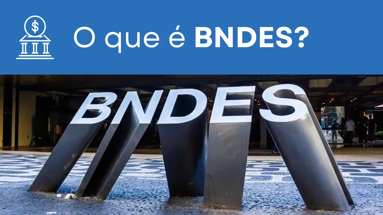Essa imagem apresenta o texto: 'O que é BNDES' e mostra o logotipo do BNDES localizado na calçada em frente ao prédio da instituição no Rio de Janeiro. O logotipo é composto pelas letras 'BNDES' esculpidas em metal na calçada.
